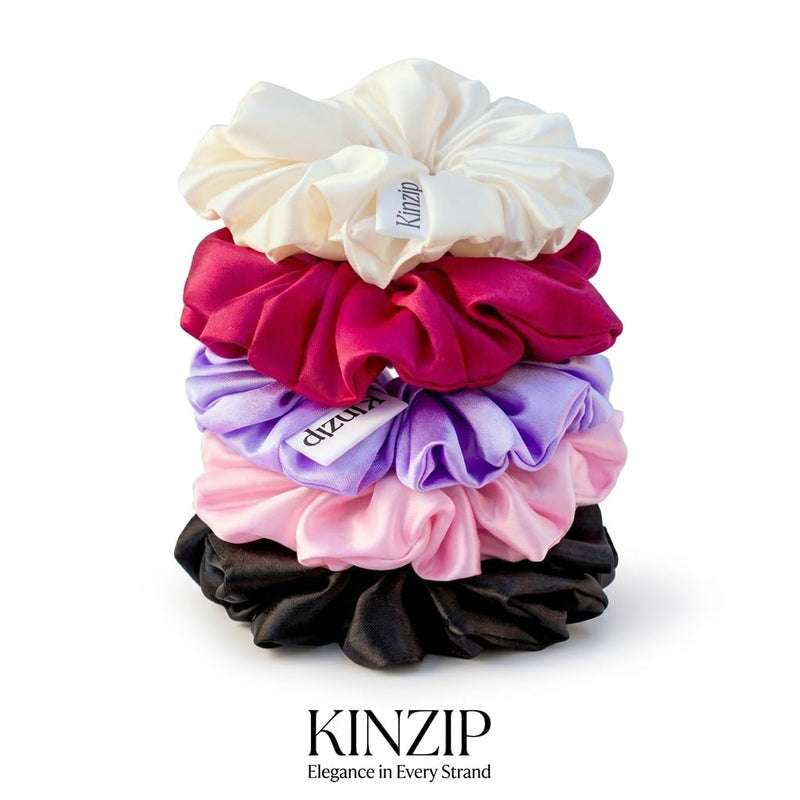 A stack of five satin hair scrunchies in cream, magenta, purple, pink, and black, with brand tags visible on each. The brand name KINZIP is printed at the bottom of the image.